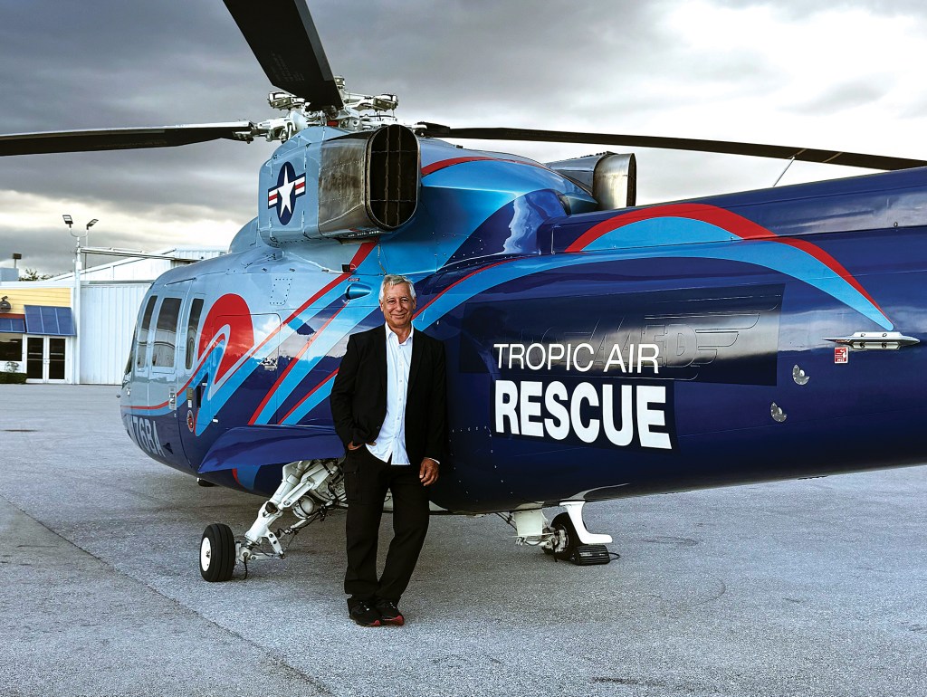 A full-length shot of founder Anthony Marinello leaning casually against a large blue rescue helicopter parked on an airport tarmac. He is smiling, wearing a dark blazer over a light button-down shirt and dark pants. The helicopter features bold red and light-blue graphics, an American star emblem near the rotor housing, and prominent white lettering on the tail boom that reads "TROPIC AIR RESCUE." A hangar building is visible in the background under an overcast, moody sky.
