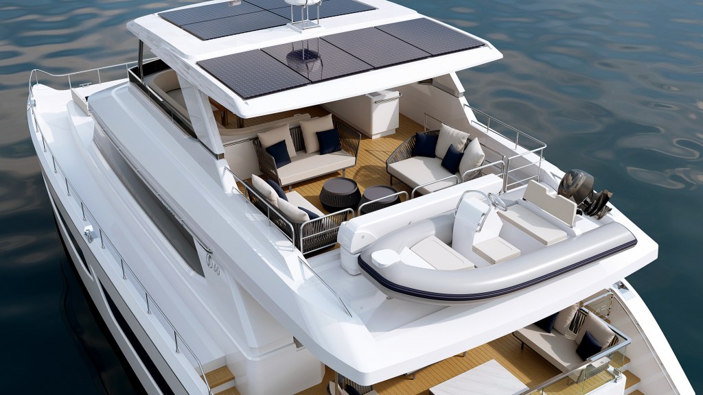 An elevated, close-up overhead shot of the flybridge and aft deck of a white luxury power catamaran sitting on calm water. The upper flybridge area features teak decking arranged with two woven outdoor sofas, a matching armchair, two round coffee tables, and a wet bar console. Six solar panels are neatly integrated into the hardtop roof structure covering the upper lounge. Mounted on the aft deck below, a grey inflatable tender boat with a center steering console rests securely on a dedicated platform.