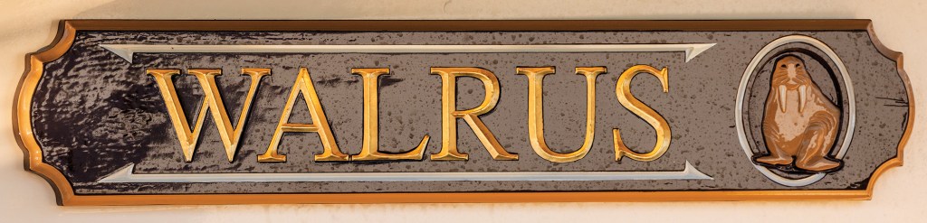 A close-up studio shot of a traditional, ornate wooden nameboard for a boat. The rectangular sign features scalloped corners and a gold-painted outer border. The name "WALRUS" is carved in large, bold, gold serif capital letters centered against a dark grey, textured background inside a silver-beveled frame. To the right of the text, a small oval carving encloses a brown illustration of a walrus with white tusks sitting upright.