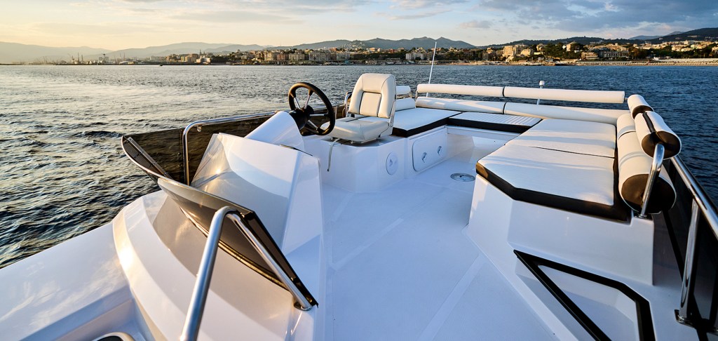 A high-angle, wide shot looking aft from the open flybridge deck of a yacht at sunset. The clean white deck features a black steering wheel and a single white helm chair on the left, alongside a large wrap-around lounge sofa with white cushions and dark trim on the right. In the background, the calm ocean stretches toward a distant coastal city skyline tucked beneath a soft evening sky.