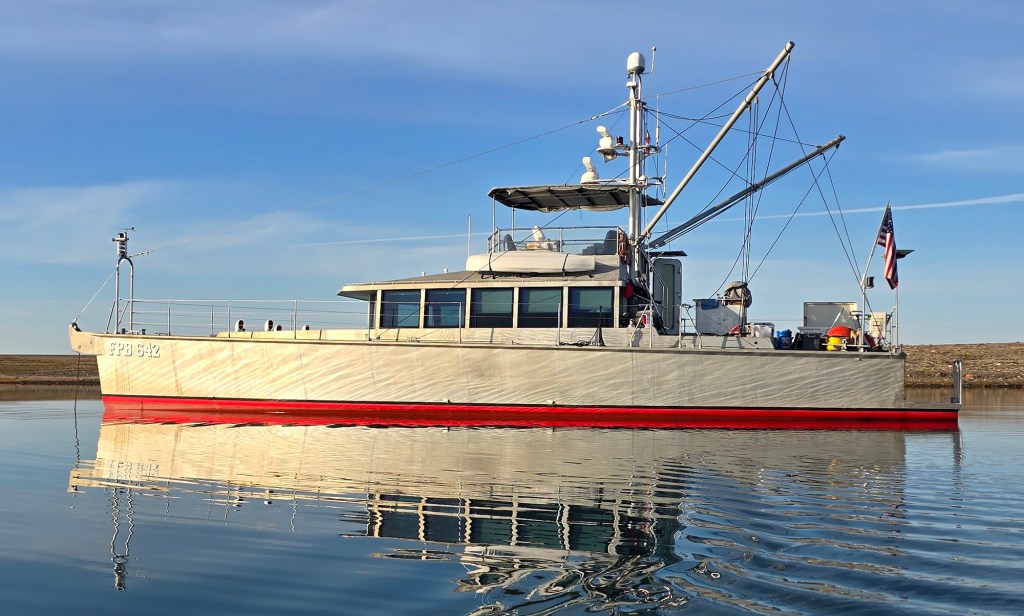 A broadside, eye-level shot of the "Sarah-Sarah," an unpainted aluminum FPB 64 long-range trawler, floating stationary on perfectly calm, glassy water that reflects the boat's shape. The rugged, military-style exploration vessel features a bright red bottom hull line contrasting against its metallic silver superstructure. The boat is designed with a long bow, a low-profile main cabin with numerous square windows, an open flybridge helm, a tall central radar mast with stabilizing outriggers, and an American flag flying from the stern under a clear blue sky.