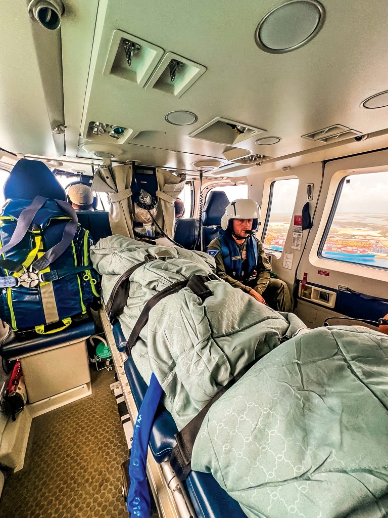 An interior shot looking down the cabin of a Tropic Air Rescue medical evacuation helicopter in flight. In the center, a medical stretcher is covered with a light green patterned blanket and secured with thick black safety straps. To the right, a flight crew member wearing a white helmet and a blue life vest sits near a cabin window that reveals a high-angle aerial view of a port facility and water below. A large blue medical response bag is strapped into a seat on the left side of the aircraft.