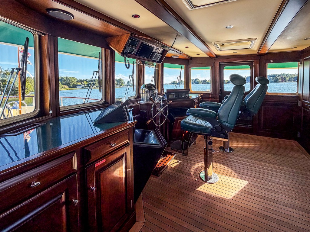 An interior view of a spacious, classic ship's wheelhouse on a yacht. The cabin features rich, dark wood cabinetry and trim, a traditional stainless steel steering wheel, and striped teak-and-holly flooring illuminated by sunlight. Two large, adjustable captain's chairs upholstered in teal leather stand on chrome pedestals facing the forward helm station. A panoramic row of large, rectangular windows with overhead green sunshades completely surrounds the bridge, offering a clear view of calm blue water and a tree-lined shoreline under a bright sky.
