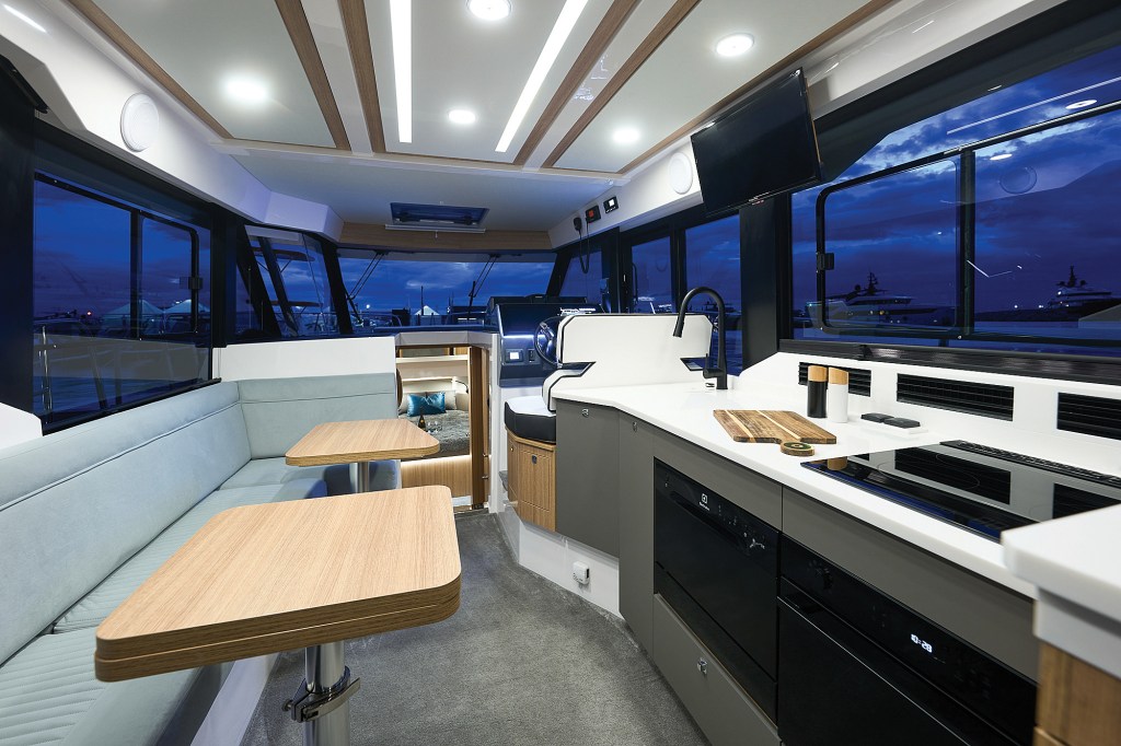 An interior view of a modern yacht salon at dusk, showcasing a bright, brightly lit cabin surrounded by large windows looking out at a deep blue twilight sky. On the left, a light blue upholstered L-shaped sofa faces two small wooden dining tables. On the right, a sleek galley features a white countertop with a built-in sink, electric cooktop, cutting board, and black appliances. A central companionway leads forward toward a glimpse of a lower stateroom bed.