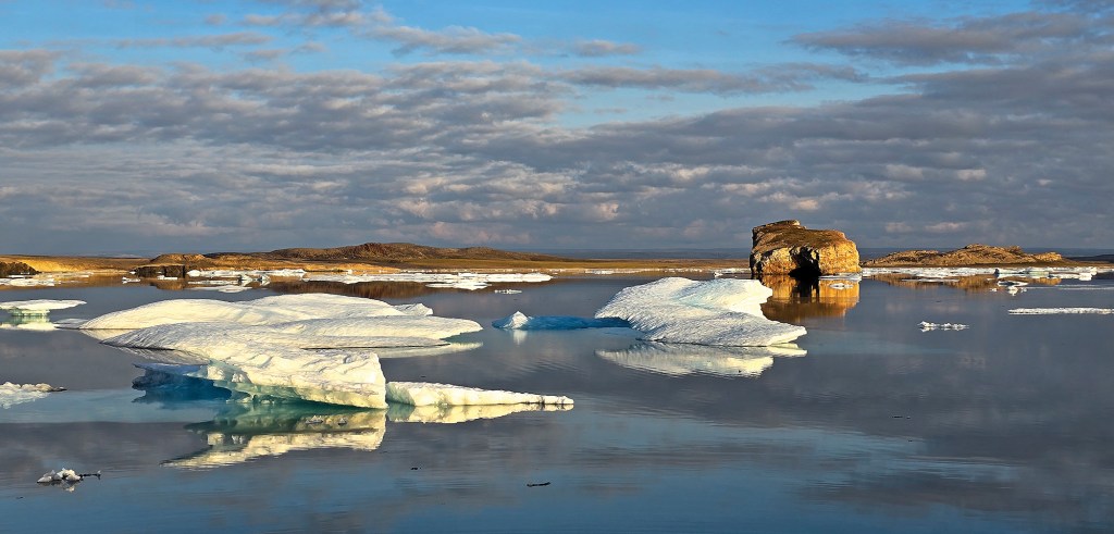 A wide, scenic landscape shot of flat sea ice floes drifting across still, mirror-like Arctic waters. In the mid-ground, low-profile white and pale blue ice sheets float near a prominent, blocky brown rock formation that stands out from the distant, barren coastline. The glassy water creates sharp, clear reflections of the ice, the small islands, and the vast sky filled with scattered, soft blue and white clouds catching the subtle light.
