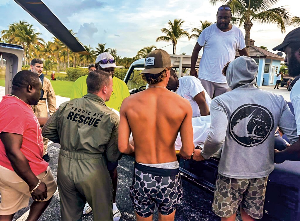 A medium, eye-level shot showing a group of people and a Tropic Air Rescue crew member carefully transferring a patient wrapped in white sheets from the back of a utility vehicle. On the left, a flight medic in a green "TROPIC AIR RESCUE" jumpsuit assists several local bystanders in holding the stretcher. A large helicopter blade is partially visible in the top left corner, cutting across a background filled with tropical palm trees under a bright sky.