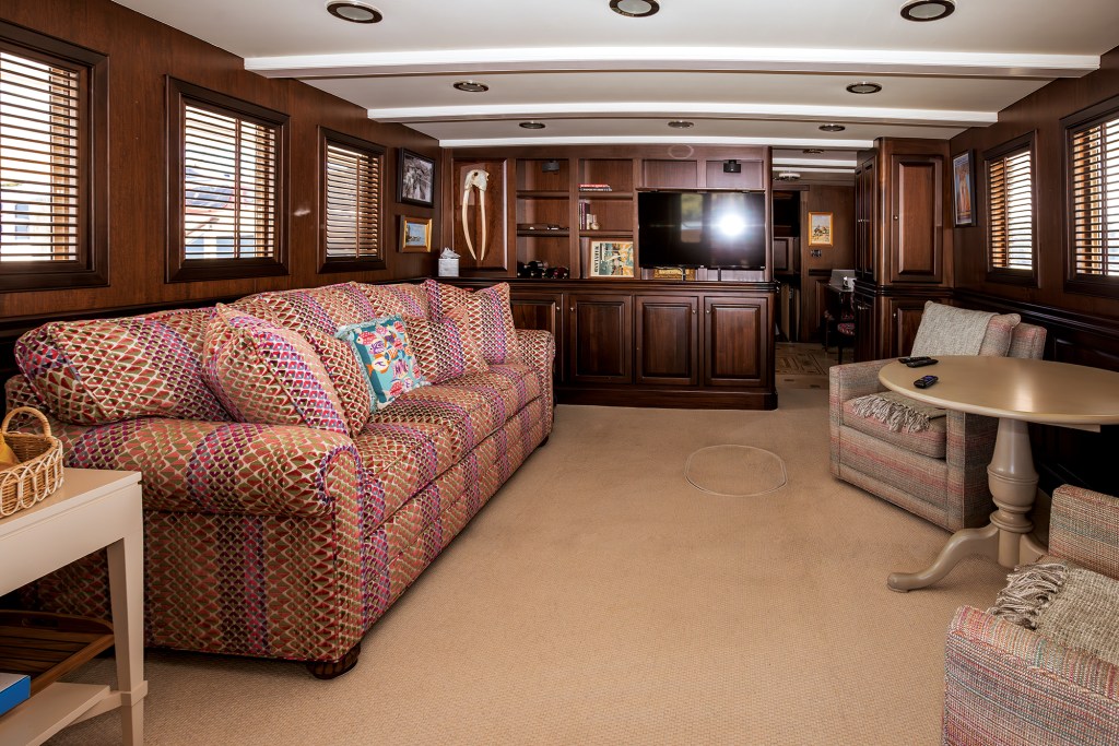 An interior view of a cozy, traditional main salon aboard a yacht. The room features warm, satin-finished dark wood paneled walls, a white beamed ceiling with recessed lighting, and beige carpeting. On the left sits a large, plush sofa upholstered in a vibrant red, pink, and green geometric patterned fabric. The opposite side of the room features two smaller fabric armchairs arranged around a circular, single-pedestal wooden table. In the background, a large flat-screen television is integrated into a custom wood entertainment center alongside built-in bookshelves, where a decorative set of walrus tusks is mounted on the wall.