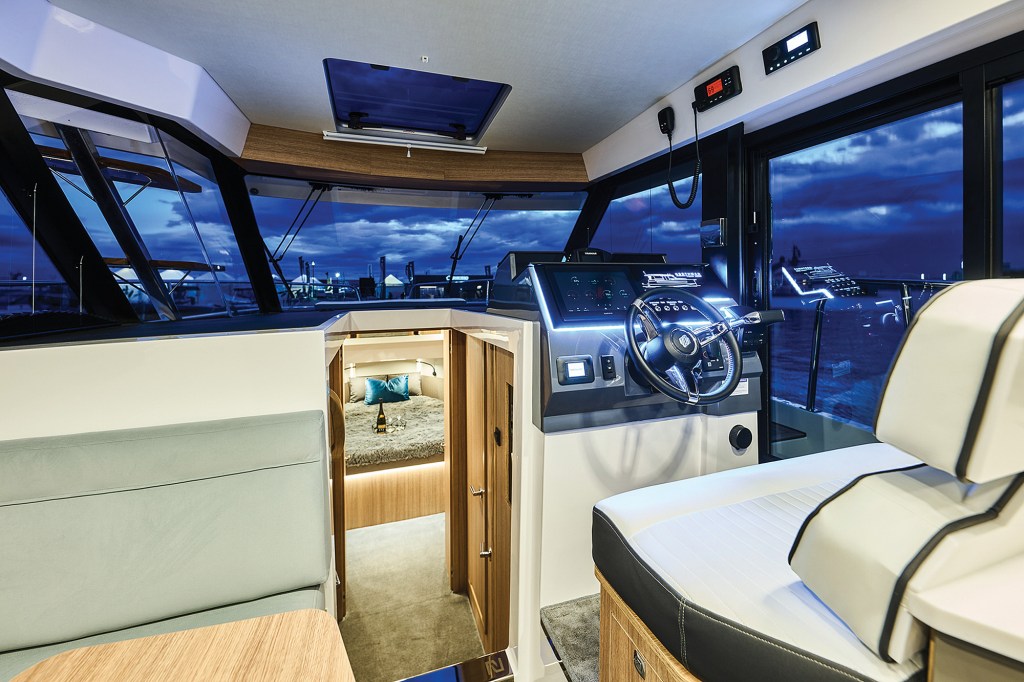 An interior view focusing on the forward helm station of a modern yacht at night. On the right, a high-tech console features a black leather-trimmed steering wheel, integrated digital touchscreens glowing with blue accent lighting, and a white upholstered captain's seat with dark piping. To the left of the helm, an open central entryway leads down into a lit forward stateroom showing a bed with a blue accent pillow. Large wrap-around windows reveal a deep blue twilight sky over an outdoor marina setting.
