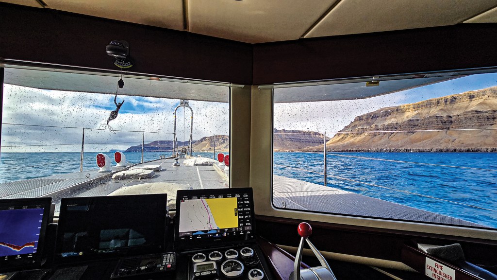 An interior view looking out through the front helm windows of a vessel navigating close to a rugged coastline. The long, industrial-style aluminum bow of the boat stretches forward, punctuated by red-capped air vents, safety rails, and a small mermaid figurine hanging near the windshield. In the lower foreground, the dashboard is equipped with multiple digital navigation displays, including a GPS chart plotter and a sonar screen showing depth data. Outside the large windows, the boat cruises through deep blue water past towering, barren brown cliffs and mountains under a partly cloudy sky.