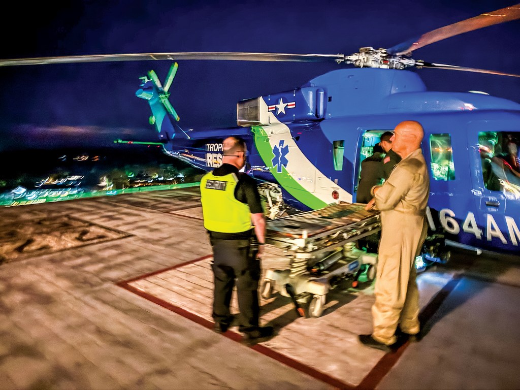 A night shot of a bright blue Tropic Air Rescue medical helicopter idling on an elevated hospital helipad. In the foreground, a medical crew member in a tan flight suit stands next to an empty wheeled stretcher alongside a security officer wearing a neon yellow safety vest. Other flight personnel work near the helicopter's open sliding side door, which features a green and white medical Star of Life emblem. In the background, city lights blur beneath a dark night sky.