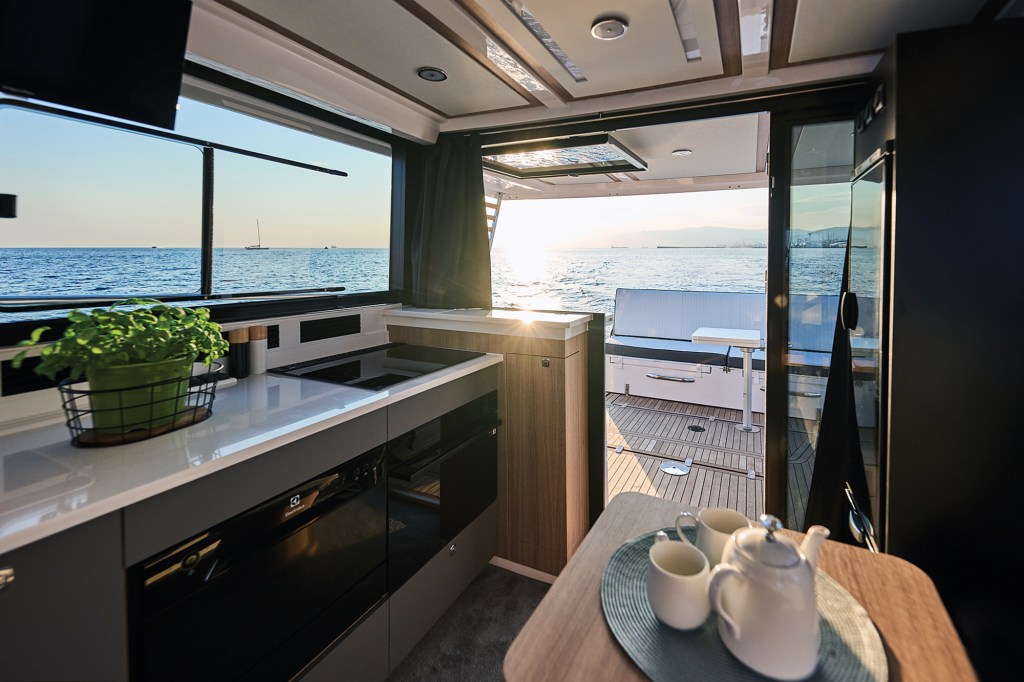 An interior view looking aft through the open glass doors of a modern yacht salon toward the cockpit deck at sunset. In the foreground, a corner of a wooden dining table holds a white ceramic teapot and mugs on a blue placemat. On the left, a sleek galley kitchen features a white countertop with a potted green plant, a black cooktop, and built-in appliances. The open glass doorway perfectly frames the sun setting over calm ocean waters, illuminating the wooden-decked aft cockpit and its white lounge seating.