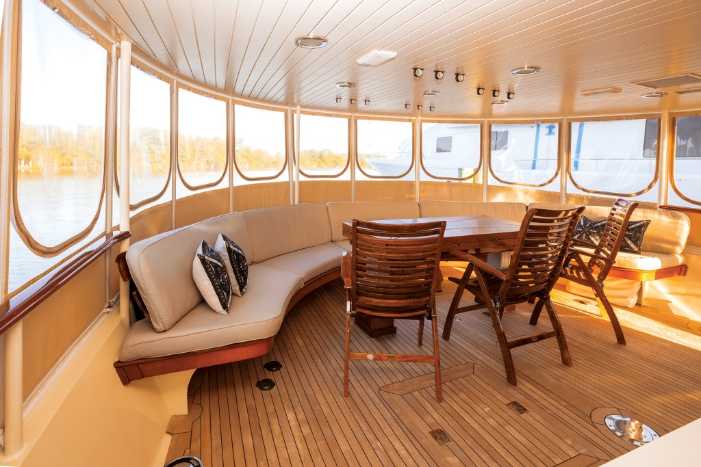 An interior view of a spacious, covered aft deck dining area on a yacht. The space features traditional teak-plank flooring and a polished wood-paneled ceiling with recessed lighting. A large, curved settee upholstered in light beige cushions wraps along the stern, accented with black-and-white patterned throw pillows. A rectangular wooden dining table sits in the center, paired with matching wooden folding chairs. The entire deck is enclosed by clear vinyl weather curtains, offering a bright view of the surrounding water and shoreline.