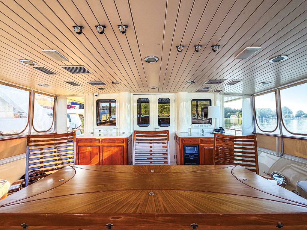 An interior view looking forward from the covered aft deck dining area of a yacht. In the foreground is a large, polished wood dining table featuring an intricate starburst veneer pattern, flanked by wooden slatted-back chairs. The background shows the cream-colored cabin superstructure with a central set of double doors and surrounding windows that look out onto the water. Polished wood cabinetry with storage and a small beverage refrigerator are built against the cabin bulkhead under a light-colored paneled ceiling fitted with recessed lights and ventilation grates.