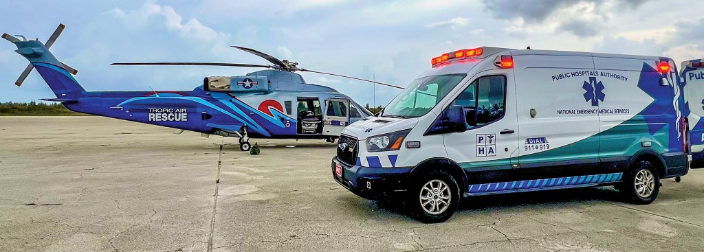 A wide, eye-level shot on a concrete tarmac showing a white and teal Public Hospitals Authority ambulance parked in the foreground with its emergency lights flashing red. In the background, a blue Tropic Air Rescue medical helicopter sits with its cabin doors open, prepared for a patient transfer. The ambulance side panel features a Star of Life medical logo and text that reads "NATIONAL EMERGENCY MEDICAL SERVICES," set against a vast, cloudy sky.