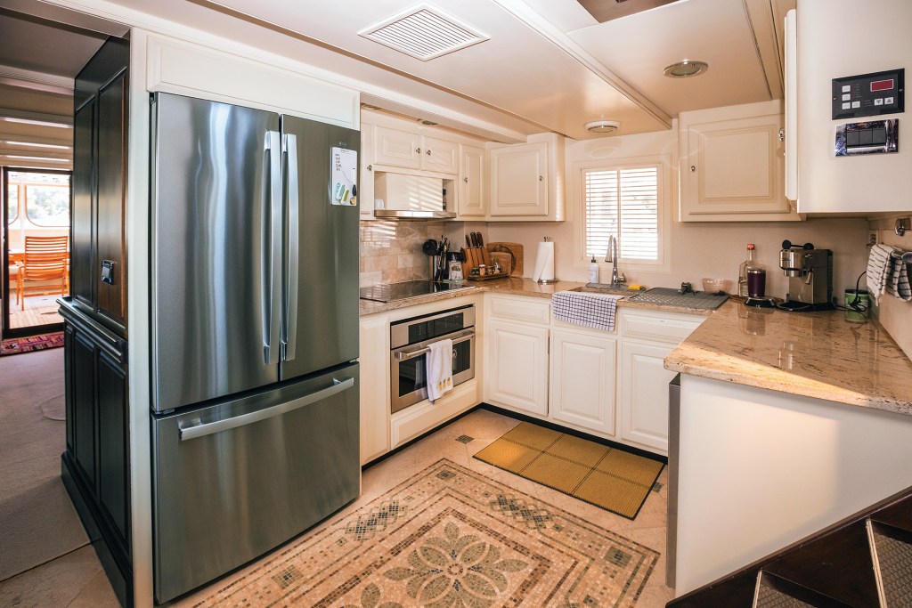 Here is the alt text copy for 05-Walrus.jpg:

Alt Text
An interior view of a bright, modern U-shaped galley kitchen aboard a yacht. On the left stands a large, French-door refrigerator in stainless steel. The main kitchen area features off-white cabinetry, speckled beige granite countertops, an undermount sink with a window behind it, and a stainless steel oven beneath a smooth cooktop. The floor is covered in light-colored tile with an intricate, decorative mosaic tile border and floral emblem centered in the space, while a doorway in the background reveals a glimpse into the salon and aft deck.