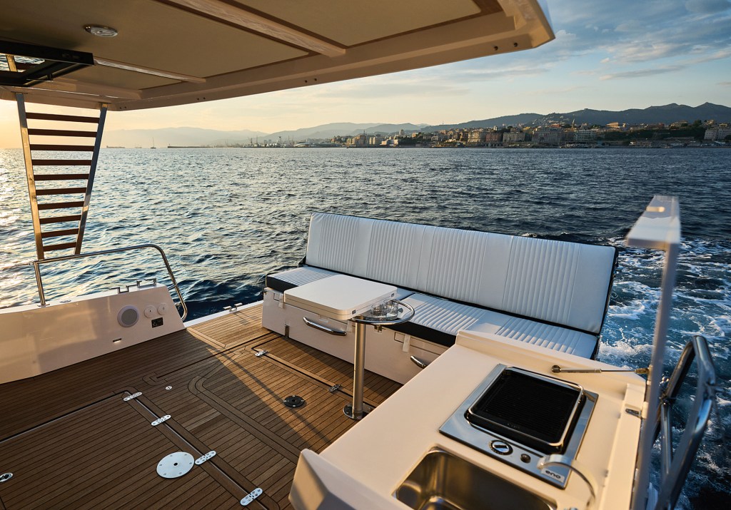 A high-angle, eye-level view of an open-air aft cockpit deck on a yacht at sunset. The deck features faux-wood teak flooring, a long bench sofa with white ribbed upholstery and dark trim, and a small pedestal table holding drinking glasses. In the right foreground, an outdoor galley unit includes a built-in stainless steel sink and an electric grill. In the background, the yacht's wake trails into calm blue ocean waters under a golden evening sky, with a distant city skyline tucked beneath coastal mountains.