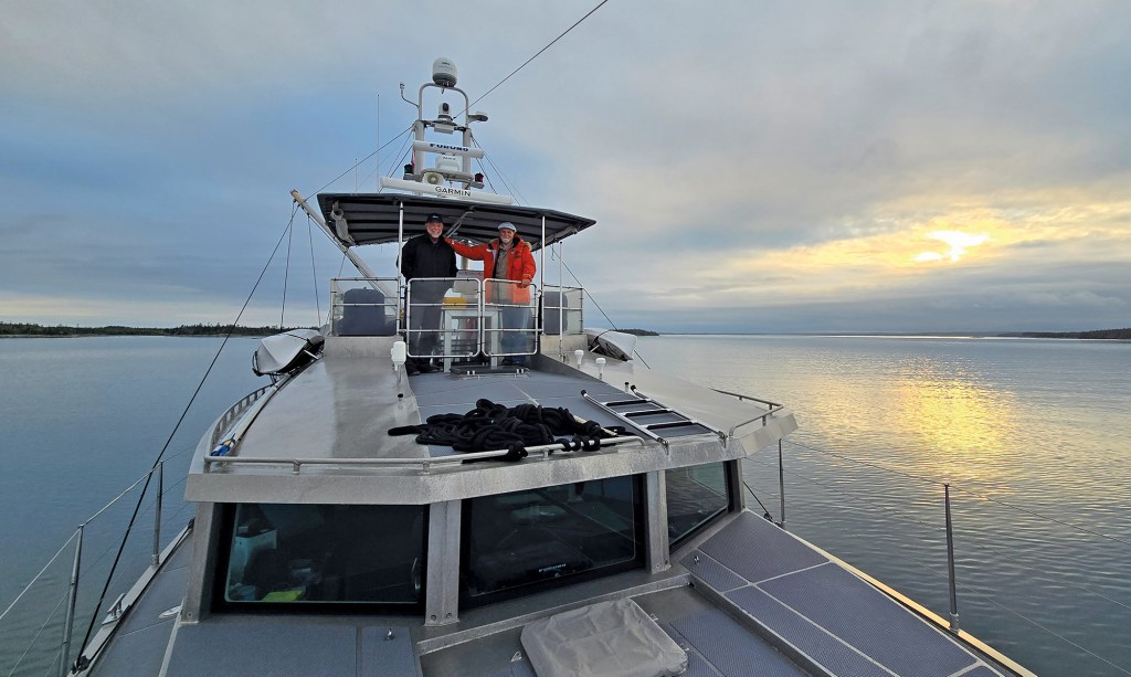 A wide, forward-facing view from the bow deck of an aluminum exploration trawler cruising through calm coastal waters at dusk. Two men stand smiling on the upper flybridge under a dark bimini canopy; the man on the left is dressed in a black jacket, while the man on the right wears a bright orange cold-weather parka with fur lining. In the foreground, the flat metallic cabin roof features a coiled black line and non-skid walking surfaces. The expansive water extends out to a flat, distant treeline under a dramatic, cloudy evening sky where the setting sun breaks through with a bright yellow glow.