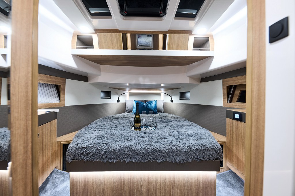 An eye-level view looking through a doorway into a modern, illuminated master stateroom cabin on a yacht. A large island bed dominates the space, made up with a plush, textured grey faux-fur blanket, grey pillows, and a vibrant teal silk accent pillow. A bottle of champagne and two flutes sit on a tray in the center of the bed. The room features warm wood paneling, indirect LED under-bed lighting, small bedside reading lamps, and a white molded ceiling with an overhead deck hatch.