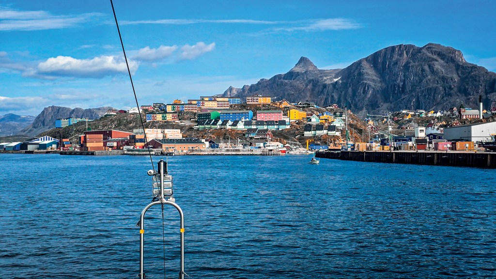 A wide, eye-level shot taken from the water approaching a colorful coastal town nestled against a dramatic mountain backdrop. In the foreground, choppy blue ocean water is visible just past a silver metal pulpit rail structure on the boat's bow. Across the harbor, rows of vibrant, multi-colored buildings in shades of red, yellow, blue, green, and pink sit clustered on a rocky hillside. The busy port area features shipping containers, industrial docks, and small boats, all framed by a jagged, rocky mountain ridge stretching high into a partly cloudy blue sky.