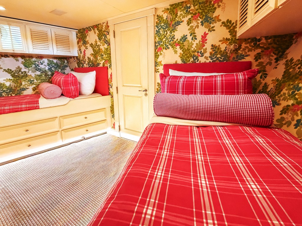 An interior view of a bright, vibrantly decorated twin stateroom cabin aboard a yacht. The room features two single built-in beds dressed in matching bright red-and-white plaid comforters, accented by pink-and-white checkered bolster pillows and solid red headboards. Underneath the left bed are built-in cream storage drawers with soft under-bed lighting reflecting onto the woven beige carpet. The walls are covered in a bold, lively floral wallpaper showcasing green leaves and red blossoms, framing a central cream-colored cabin door and white louvered overhead cabinets.
