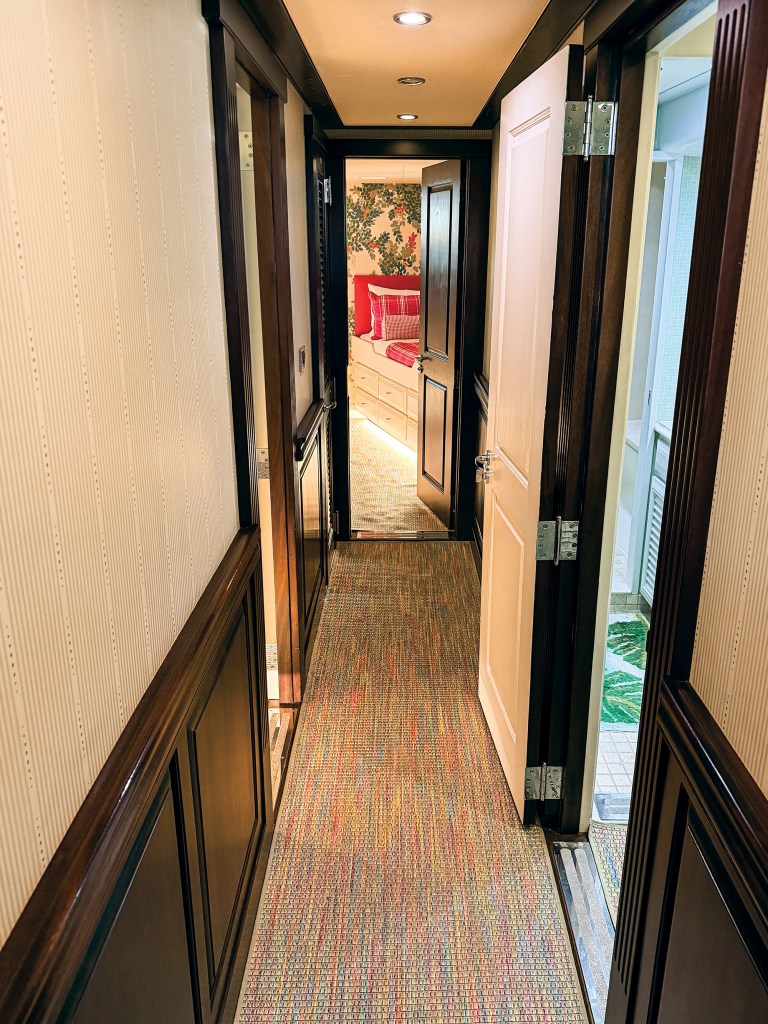 A vertical interior view looking down a narrow lower deck hallway aboard a yacht. The hallway features cream-colored, vertically striped wallpaper on the upper walls, rich dark wood wainscoting and door frames, and a colorful, textured woven runner carpet with red, orange, blue, and green hues. The ceiling has recessed spotlights. On the right, an open white door reveals a glimpse of a bathroom with a green patterned bath mat. At the far end of the corridor, an open dark wood door frames a brightly lit view into a stateroom, showcasing a bed with red plaid bedding and vibrant floral wallpaper.