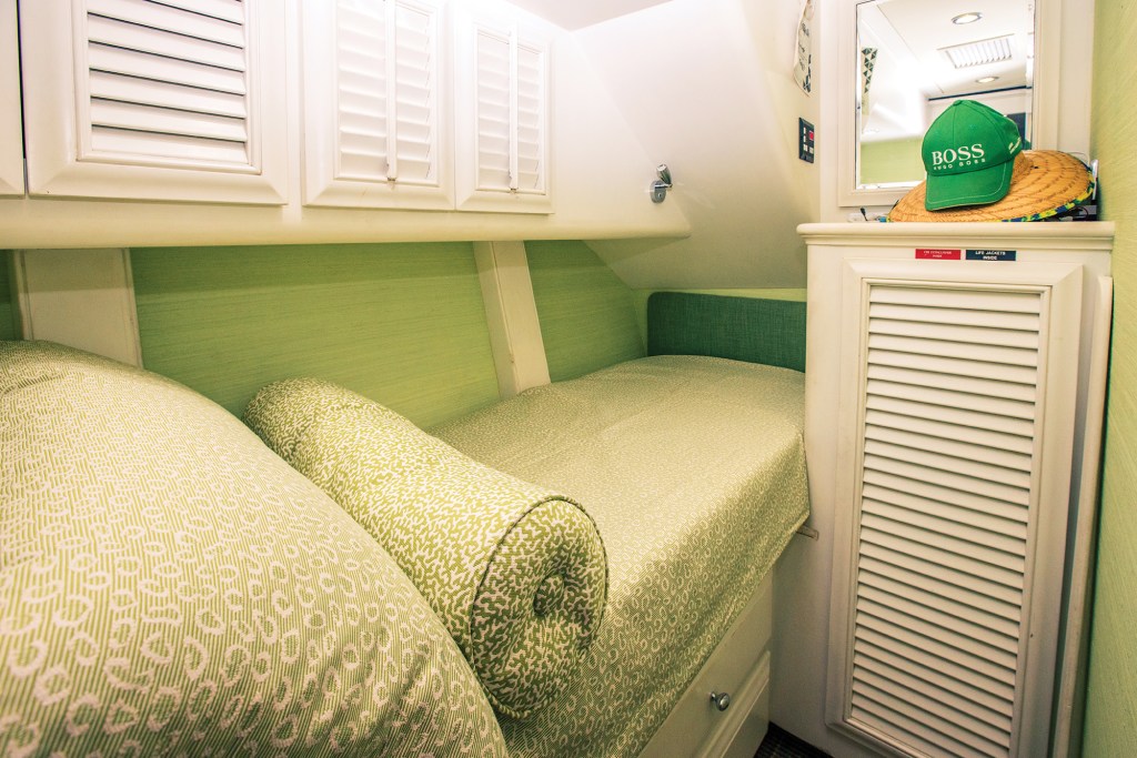An interior view of a compact, lime-green stateroom cabin aboard a yacht. A single built-in berth tucked into the hull is neatly made with a light green-and-white patterned coverlet, matching pillows, and a textured cylindrical bolster pillow. The walls are wrapped in a light green grasscloth wallpaper, accented by white built-in cabinetry featuring louvered doors above and below the bed. On top of a small storage cabinet to the right rests a woven straw hat stacked beneath a bright green "BOSS" baseball cap, situated just below a wall-mounted mirror.