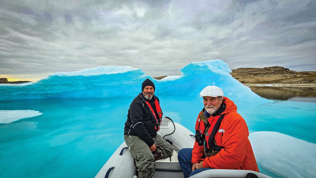 A medium, wide-angle shot from inside a white inflatable dinghy boat, featuring two smiling men cruising past a massive, vibrant neon-blue iceberg. The man on the left wears a black winter jacket, a black beanie, gray pants, and a red life vest. The man on the right wears a bright orange hooded parka, blue jeans, a white flat cap, and a black life vest with binoculars hanging around his neck. The boat glides through bright turquoise glacial water under an overcast, cloudy gray sky, with a barren, rocky brown island visible in the background on the right.