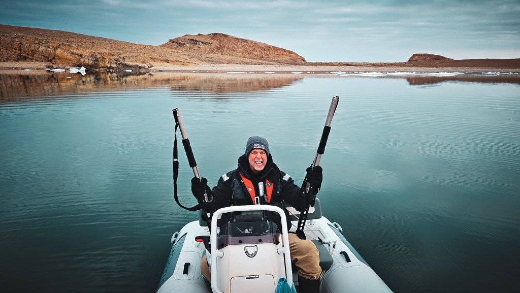 A medium, forward-facing shot of a man smiling intensely while sitting at the helm console of a white inflatable rigid-hull dinghy boat on a completely calm bay. The man wears a dark gray beanie, a heavy black winter jacket with a red life vest, khaki pants, and black gloves, holding two large silver-barreled marine poles or tools upright in each hand. The perfectly glassy, teal-tinted water reflects a barren, hilly brown landscape stretching along the distant shoreline under a pale, overcast sky.
