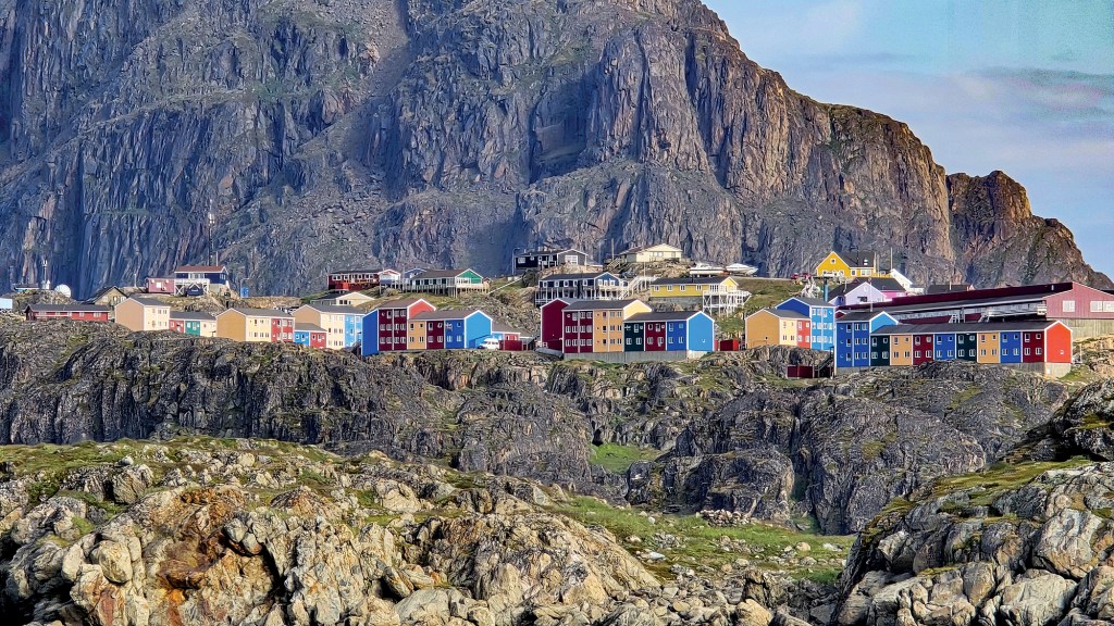 A wide, eye-level landscape shot of a coastal settlement built into a jagged, rocky terrain with a massive mountain looming directly behind it. Clusters of rectangular, Scandinavian-style apartment buildings and homes are painted in bright, vibrant colors including red, blue, yellow, and orange, standing out vividly against the rugged landscape. The foreground is composed of rough, grey-and-tan rock formations with sparse patches of green grass, leading up to the village which sits nestled at the base of a towering, sheer rock mountain face under a soft, overcast sky.