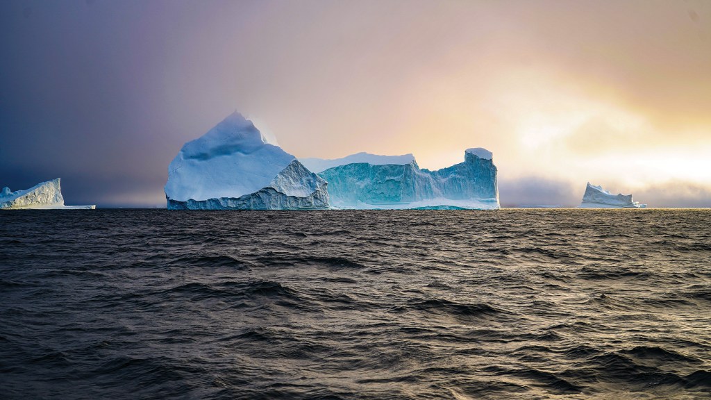 A wide, dramatic landscape shot capturing a series of large, white and light-blue icebergs floating on a dark, choppy ocean under a moody sky. The largest iceberg structure rises prominently in the center, featuring a pointed peak on the left and a long, blocky, sheer-walled plateau stretching to the right. Two smaller, isolated icebergs sit low on the horizon to the far left and right. The scene is illuminated by a striking sunset or sunrise breaking through thick clouds on the right, casting a bright, golden-orange haze over the icy water and the distant sky.