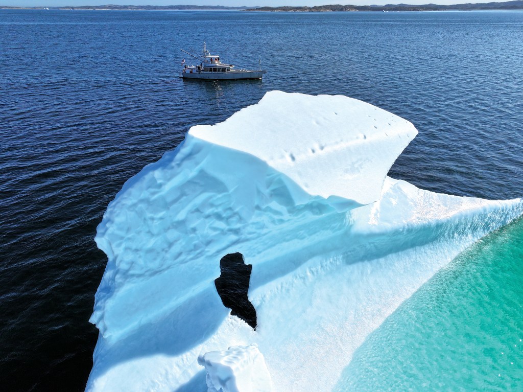 A high-angle, aerial drone shot showing a massive, sculptured white iceberg floating in a deep blue ocean, with a long-range trawler boat visible in the background. The iceberg dominates the foreground, featuring a sheer, pale blue face, a flat snowy top, a dark cavernous hole in its side, and a vibrant turquoise shelf glowing just beneath the water's surface on the right. In the upper center, the silver-and-white aluminum vessel sits safely at a distance on the calm sea, emphasizing the scale of the Arctic ice structure under a clear sky.