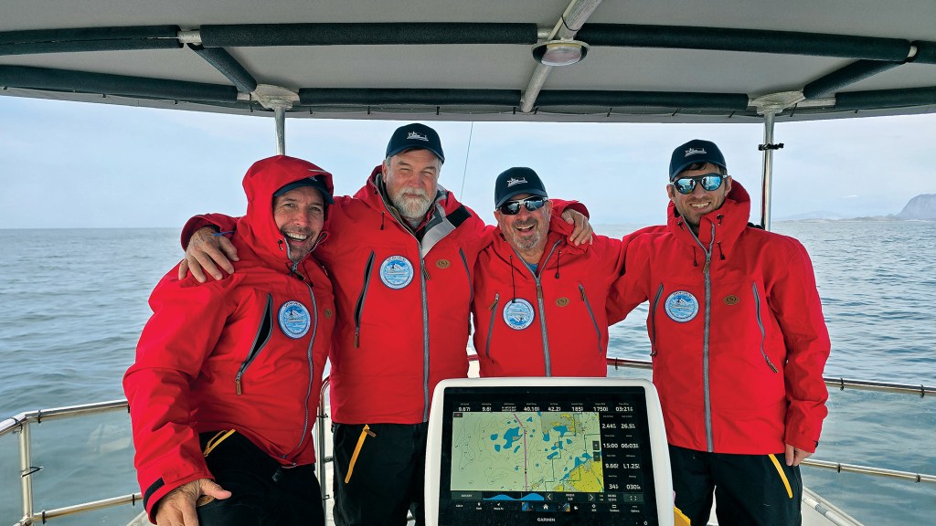 A medium, eye-level shot of four male crew members standing arm-in-arm on the open flybridge of a boat, smiling cheerfully for a group photo. All four men wear matching bright red hooded technical parkas featuring a circular maritime patch on the left chest, paired with matching dark blue baseball caps. In the lower center foreground, a large white-rimmed Garmin navigation screen displays a detailed GPS marine chart. The boat's stern railing and a grey bimini top frame the crew against a calm, grey open sea and a hazy, overcast sky.