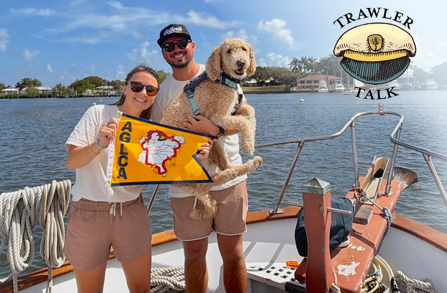 A sunny, medium shot of a smiling young man and woman posing on the bow deck of a boat with their large, fluffy golden doodle dog. The woman on the left wears a white t-shirt, brown shorts, and sunglasses while holding up a yellow triangular "AGLCA" burgee flag featuring a map graphic. The man stands behind her wearing a white t-shirt, brown shorts, a black baseball cap, and sunglasses while cradling the dog. In the top right corner, a round illustrated logo reads "TRAWLER TALK" around a captain's hat. The background shows a calm river or canal bordered by a shoreline with waterfront houses and palm trees under a blue sky.
