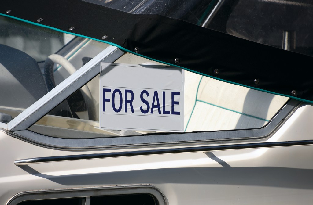 For Sale sign on a boat A close-up shot of a white boat's windshield area featuring a white rectangular "FOR SALE" sign with bold navy blue lettering taped inside the glass. A black canvas cover with metal snaps is secured tightly across the top edge of the windshield frame. Through the clear glass, a portion of the vessel's white vinyl interior seating and a white steering wheel are visible in the cockpit.