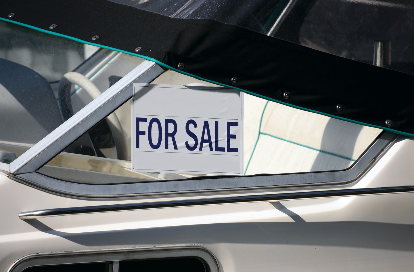 A close-up shot of a white boat's windshield area featuring a white rectangular "FOR SALE" sign with bold navy blue lettering taped inside the glass. A black canvas cover with metal snaps is secured tightly across the top edge of the windshield frame. Through the clear glass, a portion of the vessel's white vinyl interior seating and a white steering wheel are visible in the cockpit.
