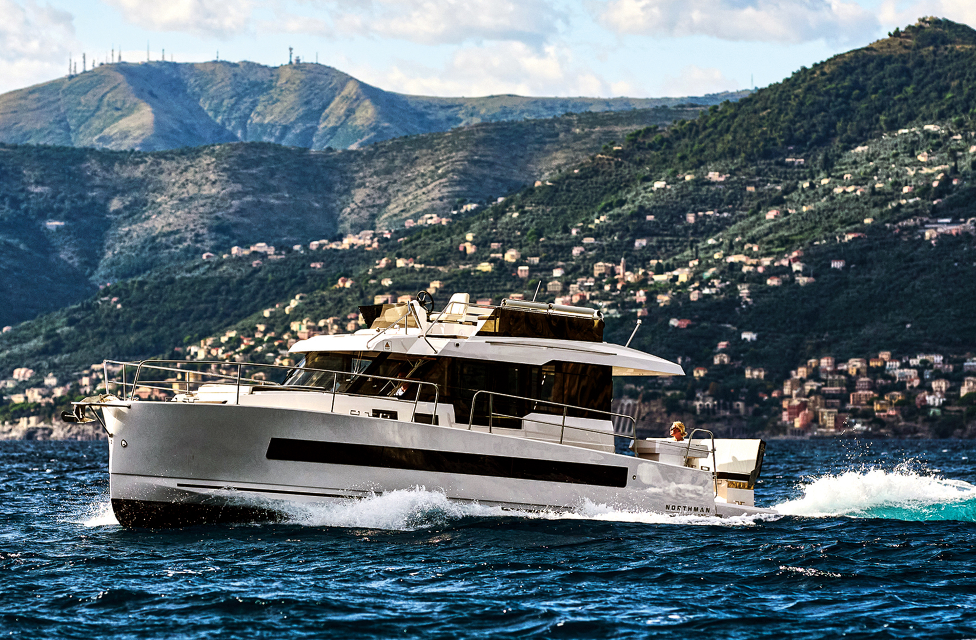 A side profile view of a modern white trawler yacht cruising through deep blue ocean water. The vessel features sleek dark hull windows, a flybridge deck, and a vertical bow that throws up a white crest of sea spray at its stem. In the background, a massive, sunlit coastal hillside rises steeply from the water, dotted with numerous residential buildings and lush green vegetation under a partially cloudy sky.