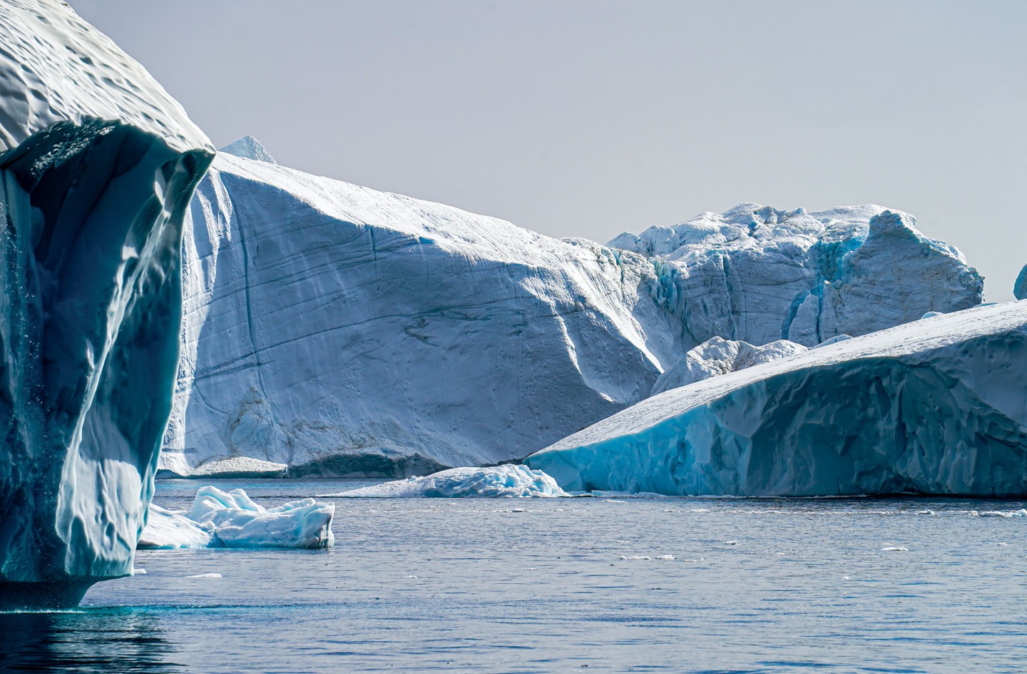 A wide, eye-level shot capturing a majestic and treacherous Arctic landscape filled with massive white and blue icebergs floating in a calm, dark blue ocean. In the immediate left foreground, a towering wall of sheer ice rises sharply, showing deep fissures and dynamic textures. Behind it, a sweeping, mountainous glacier structure dominates the background under a pale, overcast sky, while a smaller, vibrant turquoise iceberg block sits low in the water on the right, displaying bright, light-refracting edges.