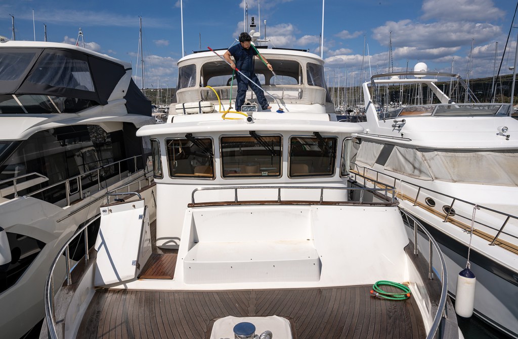 Professional worker cleaning and maintaining a luxury yacht at a A high-angle, eye-level shot looking toward the bow cabin of a large white trawler yacht docked in a marina. A worker in dark blue clothes stands atop the cabin roof structure, using a long-handled brush to scrub the fiberglass surface next to a yellow water hose. The boat features a forward-slanted windshield with multiple wipers, a dark wood-planked foredeck, and stainless steel railings. Other luxury yachts are closely docked on either side under a bright, partly cloudy blue sky.