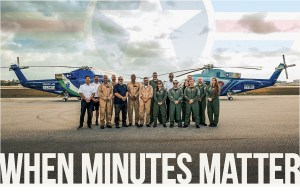 A wide shot of the Tropic Air Rescue team standing together on an airport tarmac between two large blue rescue helicopters. The flight crew, pilots, and medical personnel wear a mix of green flight suits, tan jumpsuits, and polo shirts. In the background, a cloudy sky features a large, semi-transparent watermark logo of a star with wings. Bold, white capital letters overlay the bottom of the image, reading "WHEN MINUTES MATTER."