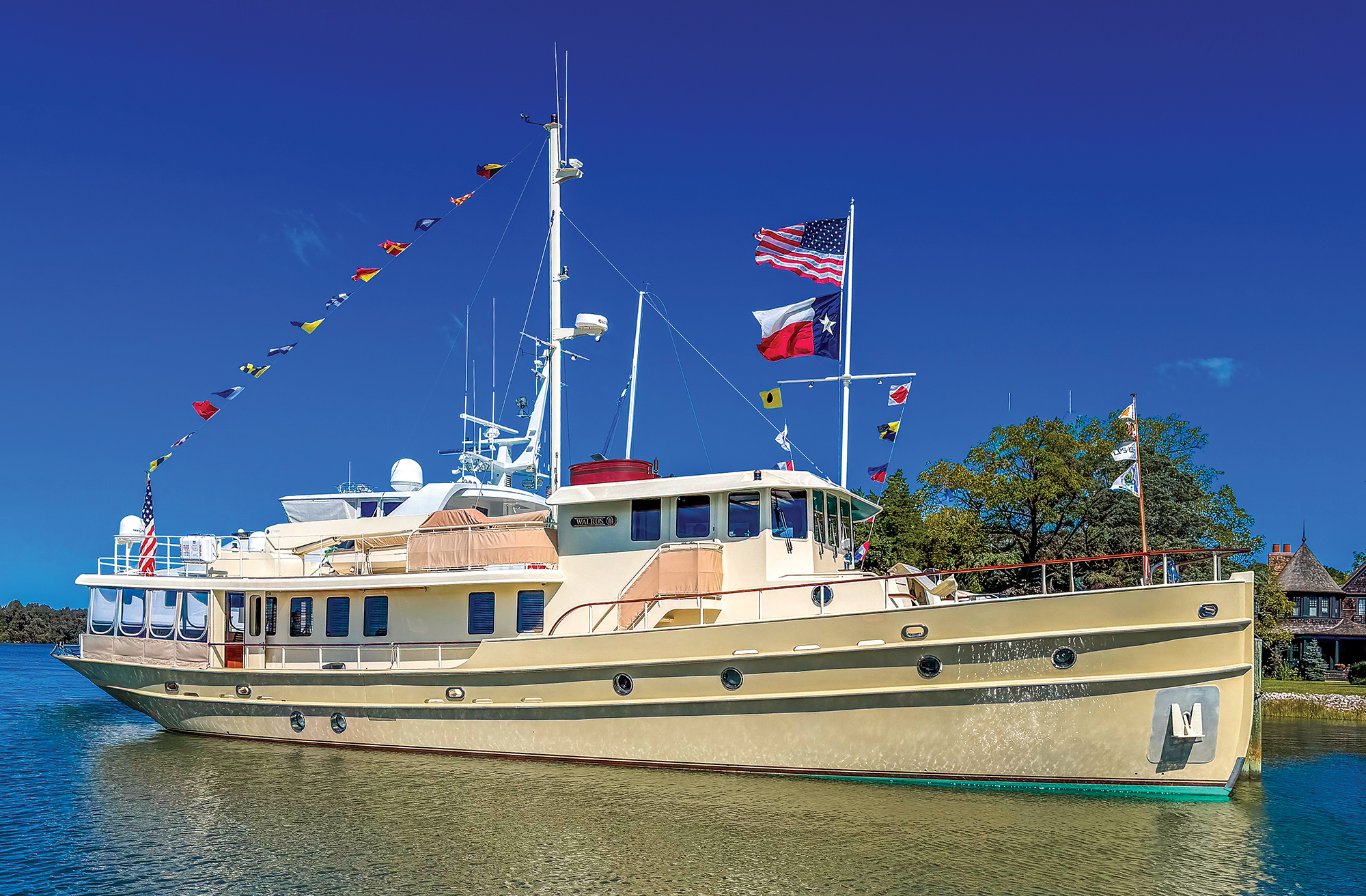 A wide, sunny shot of a large, classic expedition-style motor yacht named "Walrus" anchored in calm, shimmering water. The vessel features a cream-colored hull with circular portholes and a multi-level superstructure. It is heavily dressed in festive maritime signaling flags strung from the bow to the tall white main mast. At the stern, both an American flag and a Texas state flag fly prominently from a secondary flagpole. The background features a green, tree-lined shoreline with a glimpse of a waterfront house under a clear, vibrant blue sky.