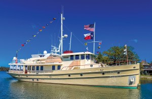 A wide, sunny shot of a large, classic expedition-style motor yacht named "Walrus" anchored in calm, shimmering water. The vessel features a cream-colored hull with circular portholes and a multi-level superstructure. It is heavily dressed in festive maritime signaling flags strung from the bow to the tall white main mast. At the stern, both an American flag and a Texas state flag fly prominently from a secondary flagpole. The background features a green, tree-lined shoreline with a glimpse of a waterfront house under a clear, vibrant blue sky.