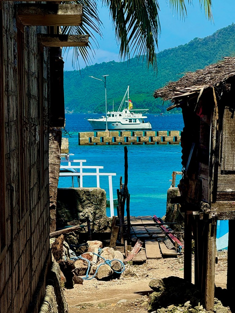 A view framed by rustic waterfront buildings showing a white Nordhavn expedition yacht anchored beyond a stone pier in bright turquoise tropical waters.