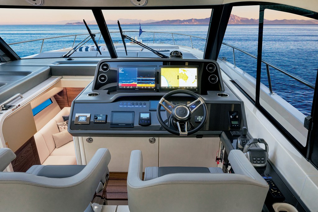 A view looking forward from inside the main cabin helm station of a modern luxury yacht, looking out across the bow toward calm open ocean and distant mountains.