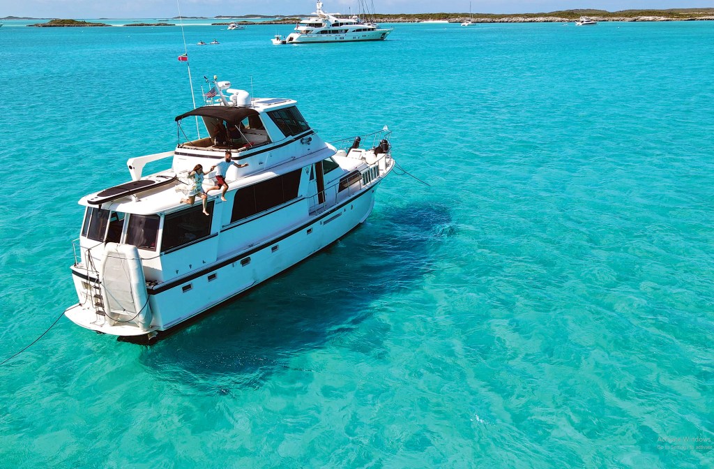 A man and a woman holding hands as they jump off the upper deck of a white motor yacht into clear, turquoise tropical water.