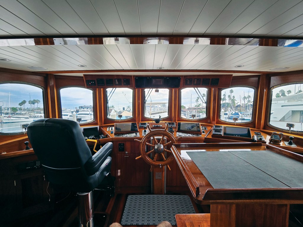 An interior view looking forward inside the spacious, wood-paneled pilothouse of the trawler Paul Johansen. The bridge features fine varnished woodwork throughout, a white slatted ceiling, and an expansive curved array of windows looking out at a marina under a cloudy sky. In the center sits a large, classic wooden ships steering wheel, flanked by a plush black leather captain’s helm chair on the left and a large wooden navigation plotting table with a dark grey mat on the right. Modern electronic navigation displays and marine instruments are integrated seamlessly into the wooden dash console.