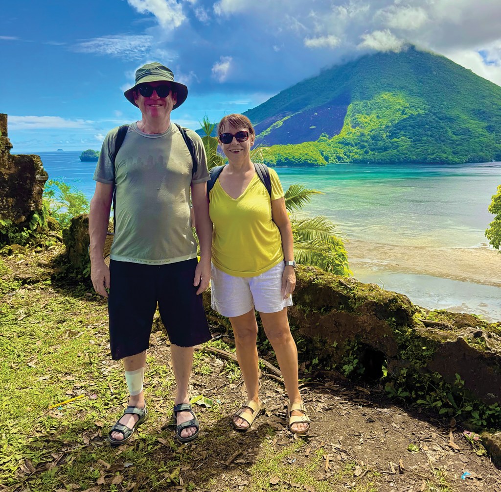 A man and a woman in sun hats and casual hiking gear smile together on a scenic overlook, with a lush volcanic mountain, clear turquoise water, and tropical islands in the background.