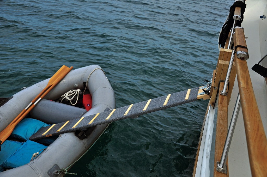 A high-angle shot of a custom pet boarding ramp covered in grey carpet with wooden traction steps extending from a yacht's side boarding gate down into an inflatable dinghy floating on open water.