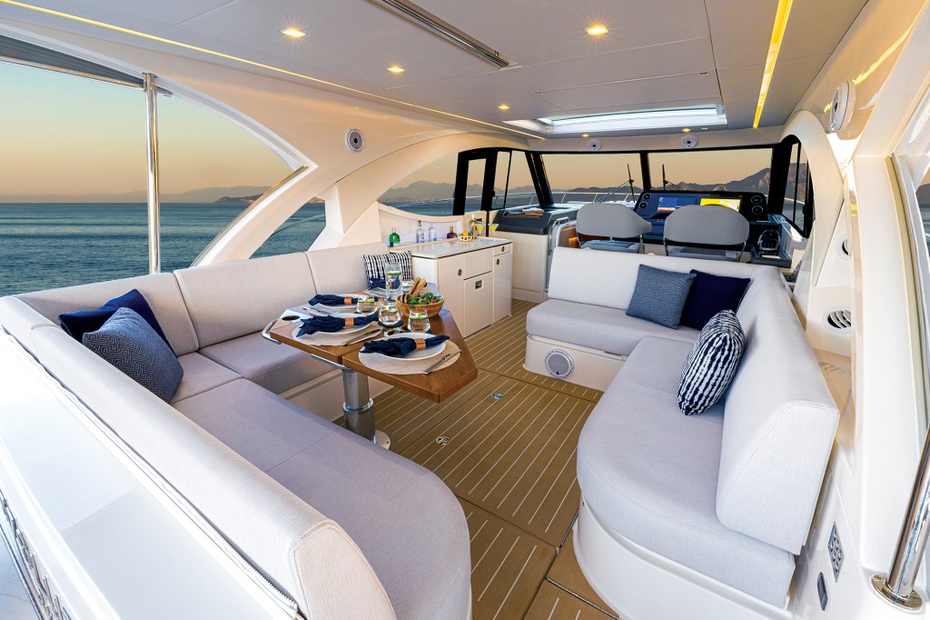 A wide-angle interior view of the spacious, open-air upper lounge and sports deck on a luxury yacht, set for dining against a sunset ocean backdrop.
