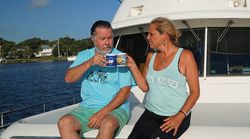 A man and a woman sitting on the deck of a boat clinking coffee mugs together on a sunny day.