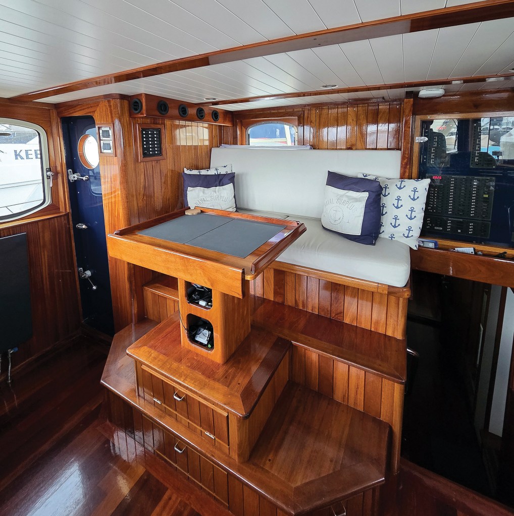 An interior shot looking toward the elevated watch berth and settee area inside the finely varnished wooden pilothouse of the trawler Paul Johansen. Wooden steps with built-in storage drawers lead up to a white cushioned bench seat decorated with nautical anchor and "Naval Surplus" accent pillows. In front of the seating area, a custom wooden navigation table with a dark grey insert sits on a multi-tiered wooden platform, which also houses electronics in open cubbies. The rich wood paneled walls, glossy wood flooring, and white slatted ceiling emphasize the boat's premium craftsmanship, with a glass-enclosed electrical control panel visible on the far right.